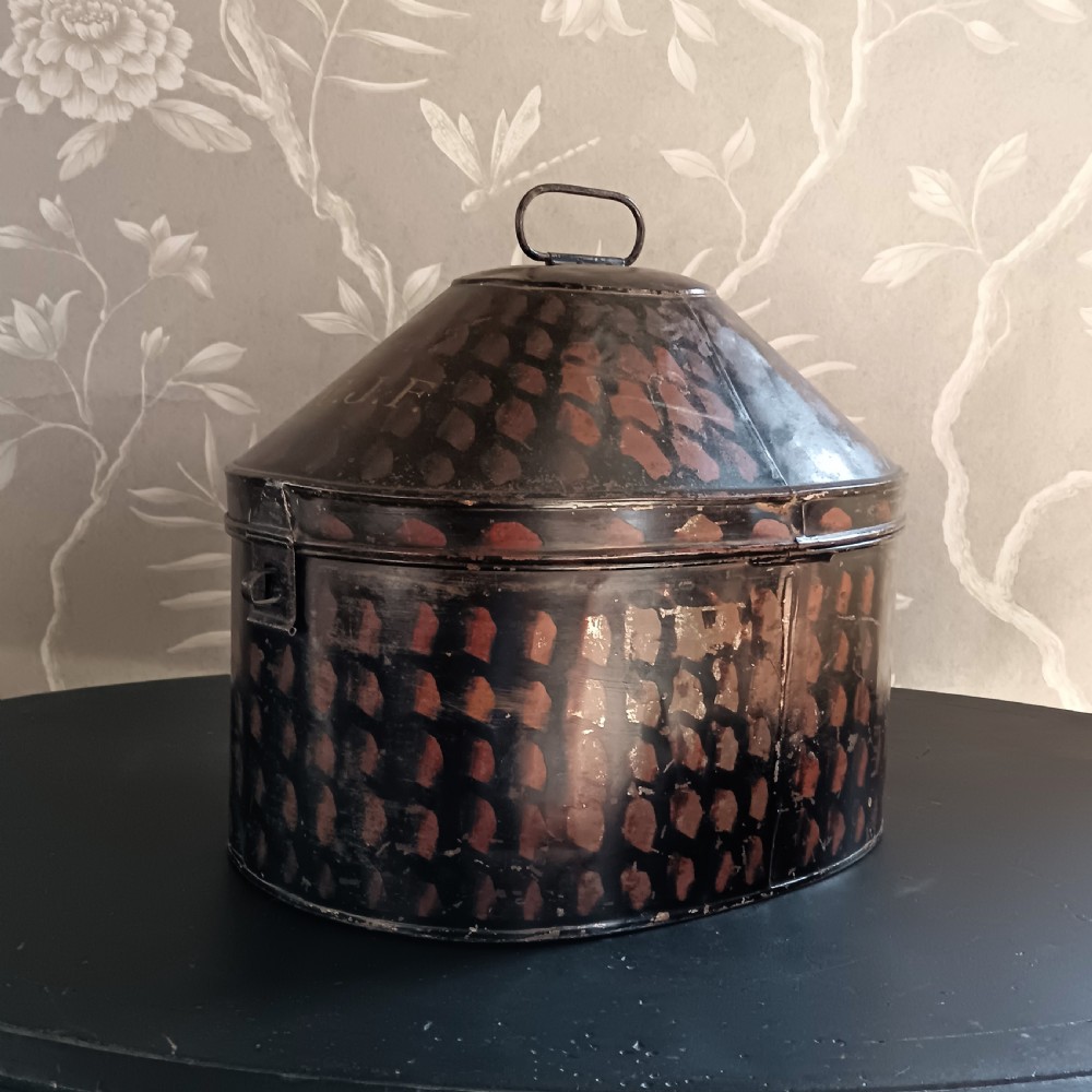 A dark painted antique tin hat box with a domed lid, top carrying handle, and hand-painted pattern.