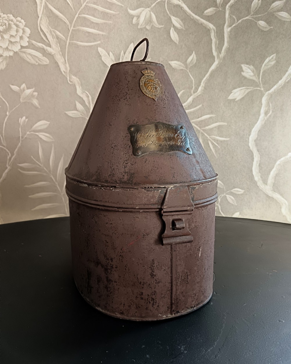 A tall, tapered antique metal hat box made from dark brown toleware, featuring a brass maker badge and an engraved brass nameplate on the front.
