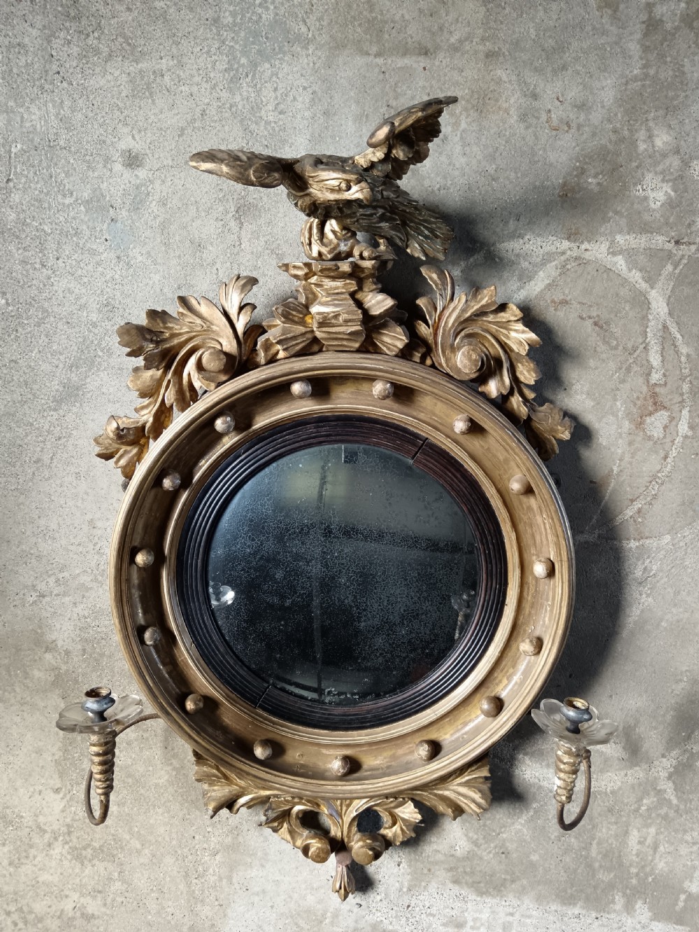 A circular Regency style giltwood convex wall mirror topped with a carved eagle cresting and featuring twin candle sconces.