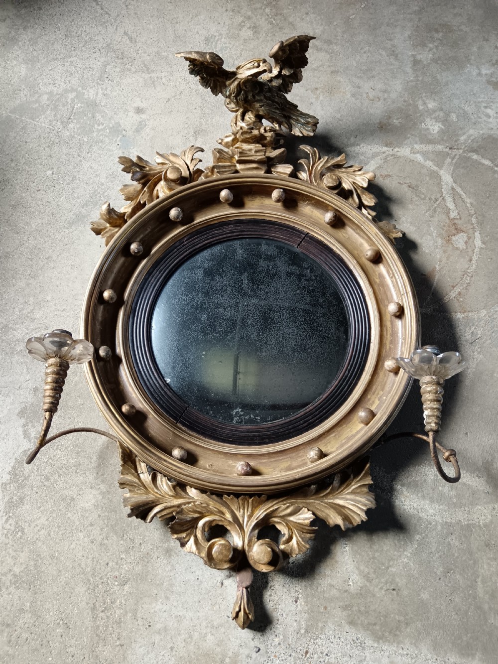 A circular Regency style giltwood convex wall mirror topped with a carved eagle cresting and featuring twin candle sconces. View II