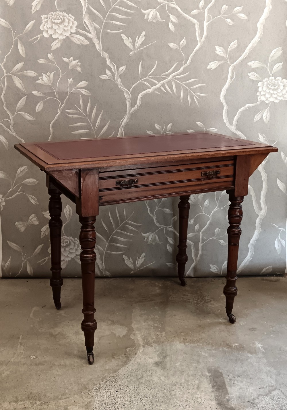 An Arts and Crafts mahogany writing desk featuring a brown leather inset top, a single wide drawer with metal handles, turned legs, and brass castors. View V