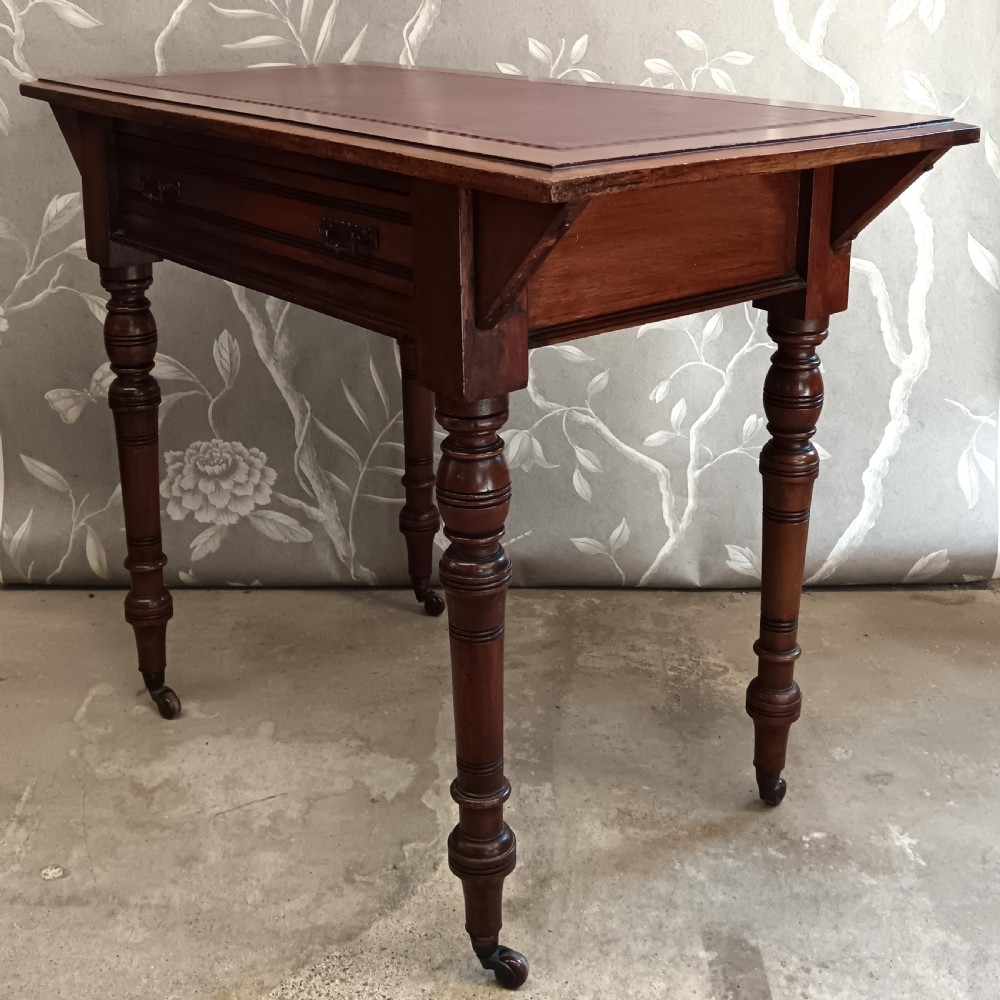 An Arts and Crafts mahogany writing desk featuring a brown leather inset top, a single wide drawer with metal handles, turned legs, and brass castors. View VI