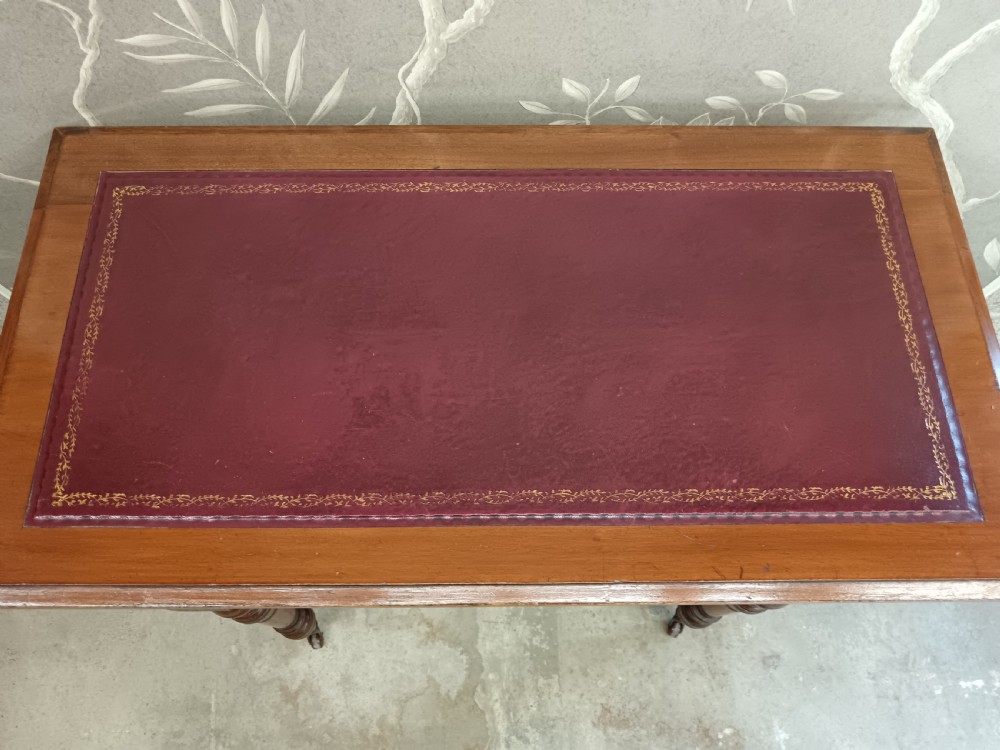 An Arts and Crafts mahogany writing desk featuring a brown leather inset top, a single wide drawer with metal handles, turned legs, and brass castors. View X