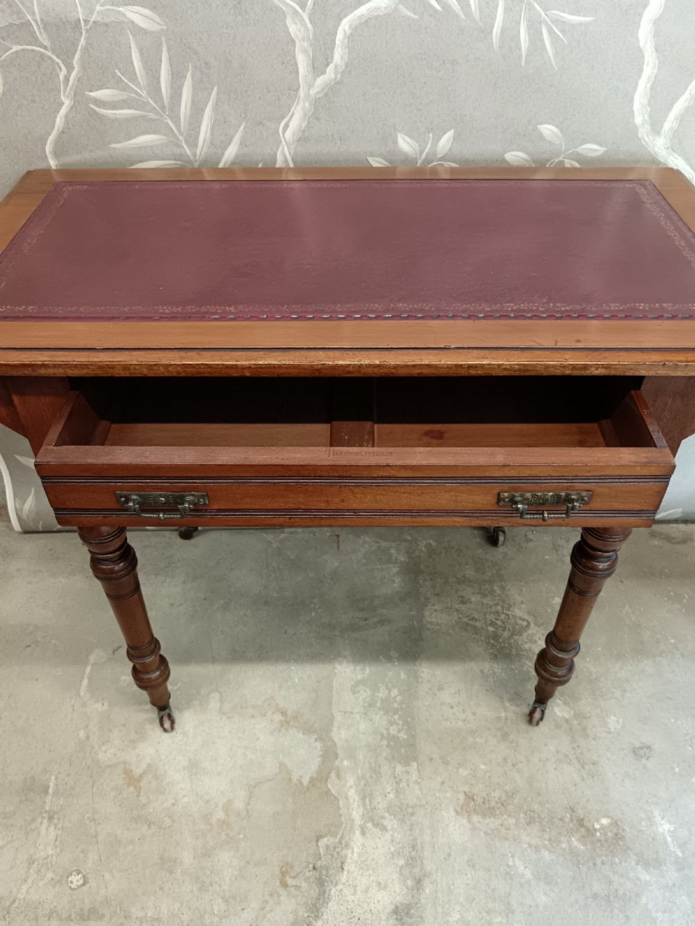 An Arts and Crafts mahogany writing desk featuring a brown leather inset top, a single wide drawer with metal handles, turned legs, and brass castors. View XI