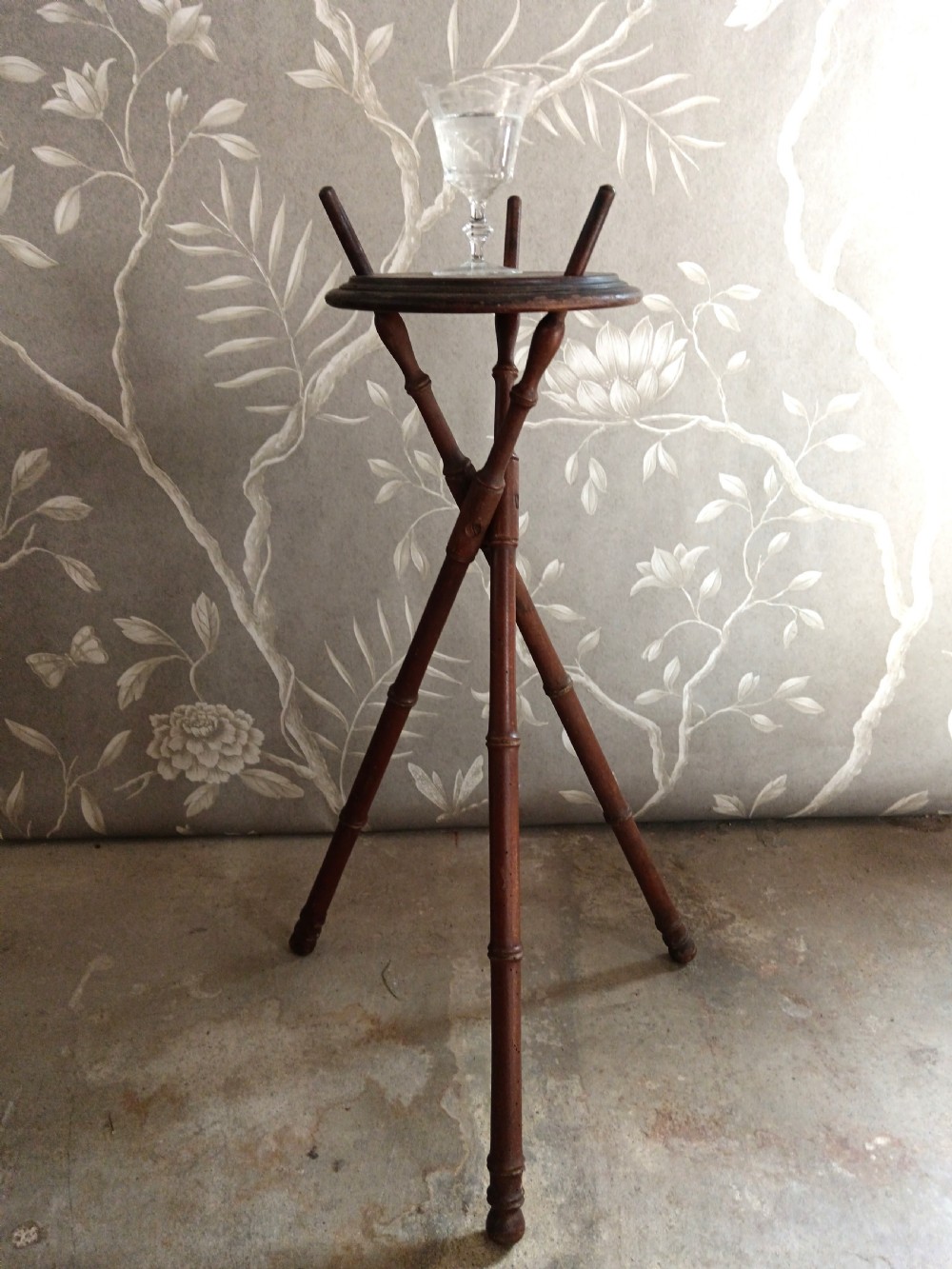 A dark brown wooden campaign washbowl stand featuring turned faux-bamboo tripod legs and vertical pegs around a circular top.