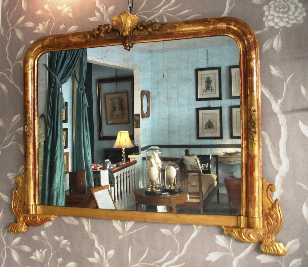 An antique gilded wood overmantel mirror featuring rounded top corners, a central carved shell crest, and scroll bracket feet.