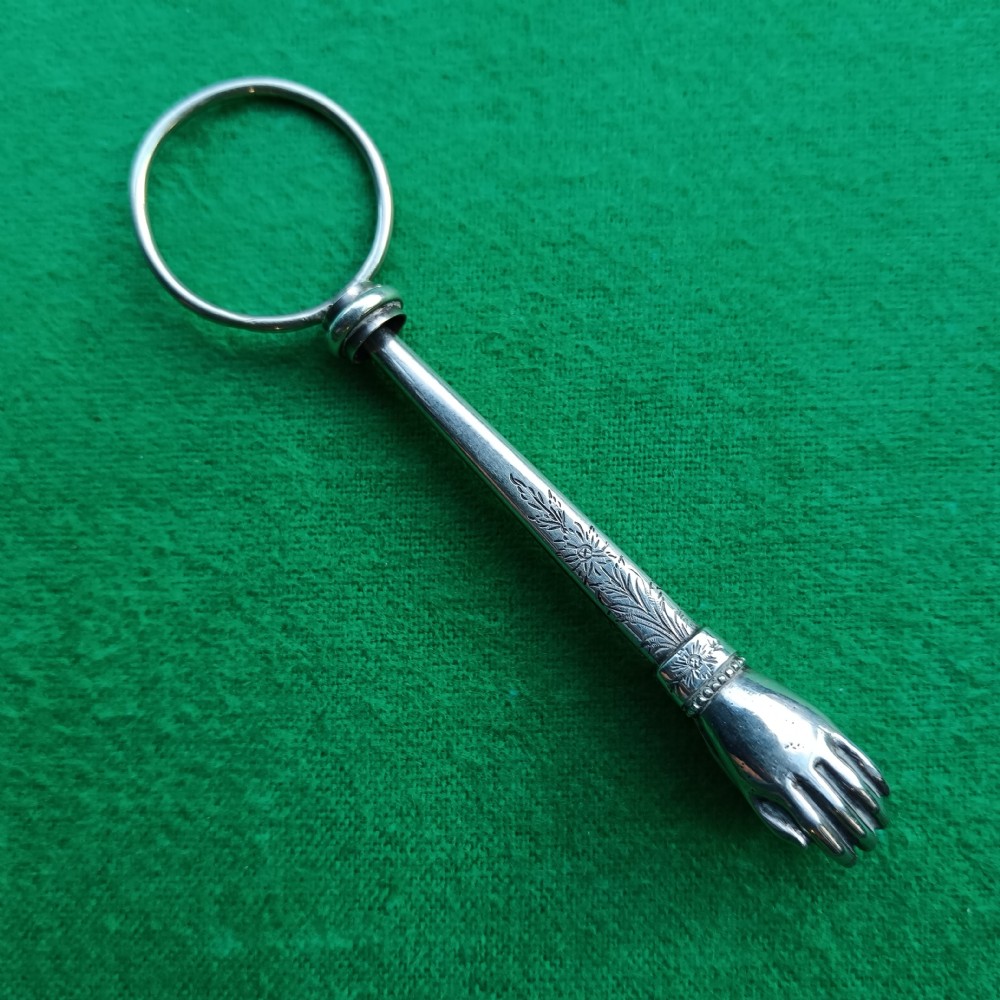 An antique Victorian silver skirt lifter featuring a top ring, an engraved shaft, and a hand-shaped terminal clip.