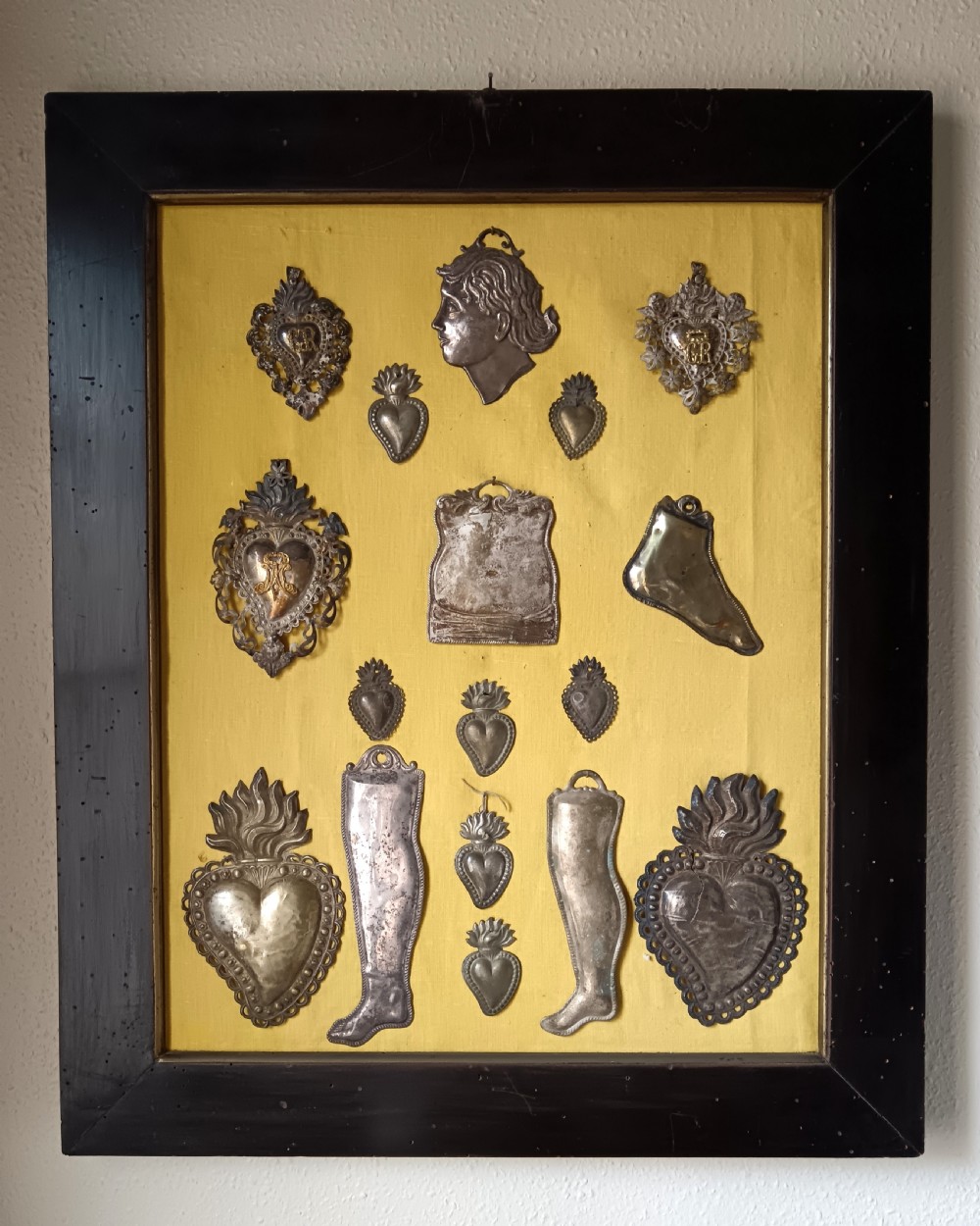 Seventeen silver-coloured metal ex-voto tokens arranged on yellow fabric inside a dark wooden frame.