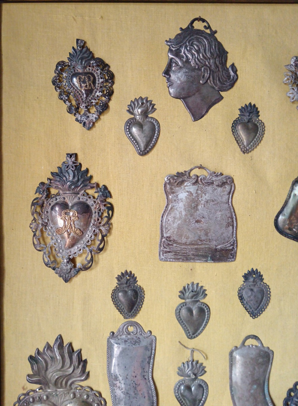 Seventeen silver-coloured metal ex-voto tokens arranged on yellow fabric inside a dark wooden frame. View II