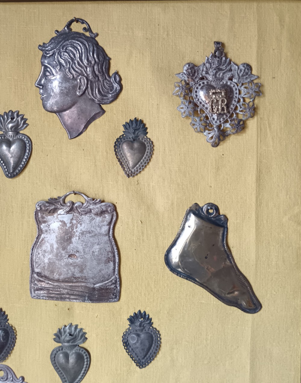 Seventeen silver-coloured metal ex-voto tokens arranged on yellow fabric inside a dark wooden frame. View III