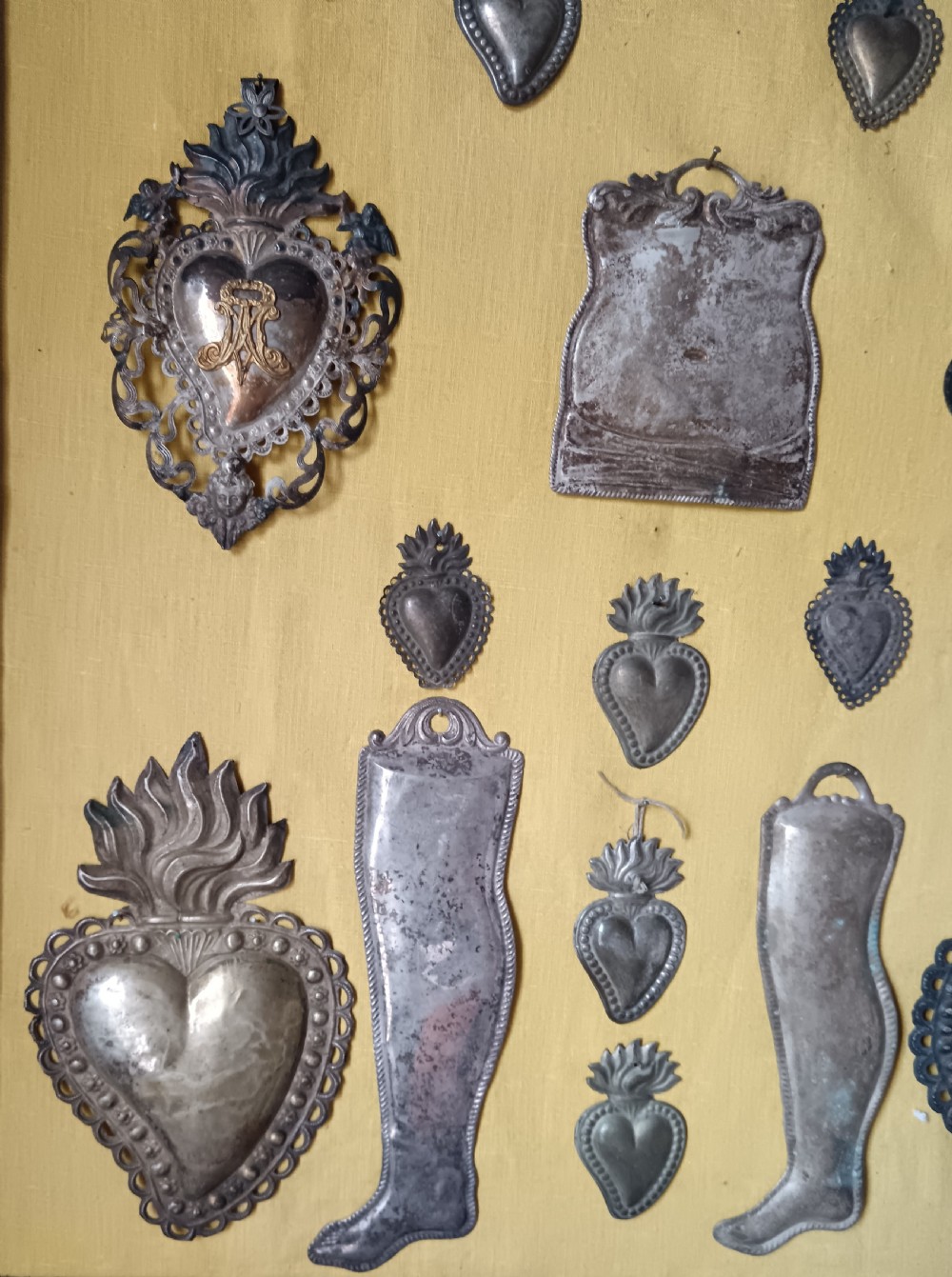 Seventeen silver-coloured metal ex-voto tokens arranged on yellow fabric inside a dark wooden frame. View V
