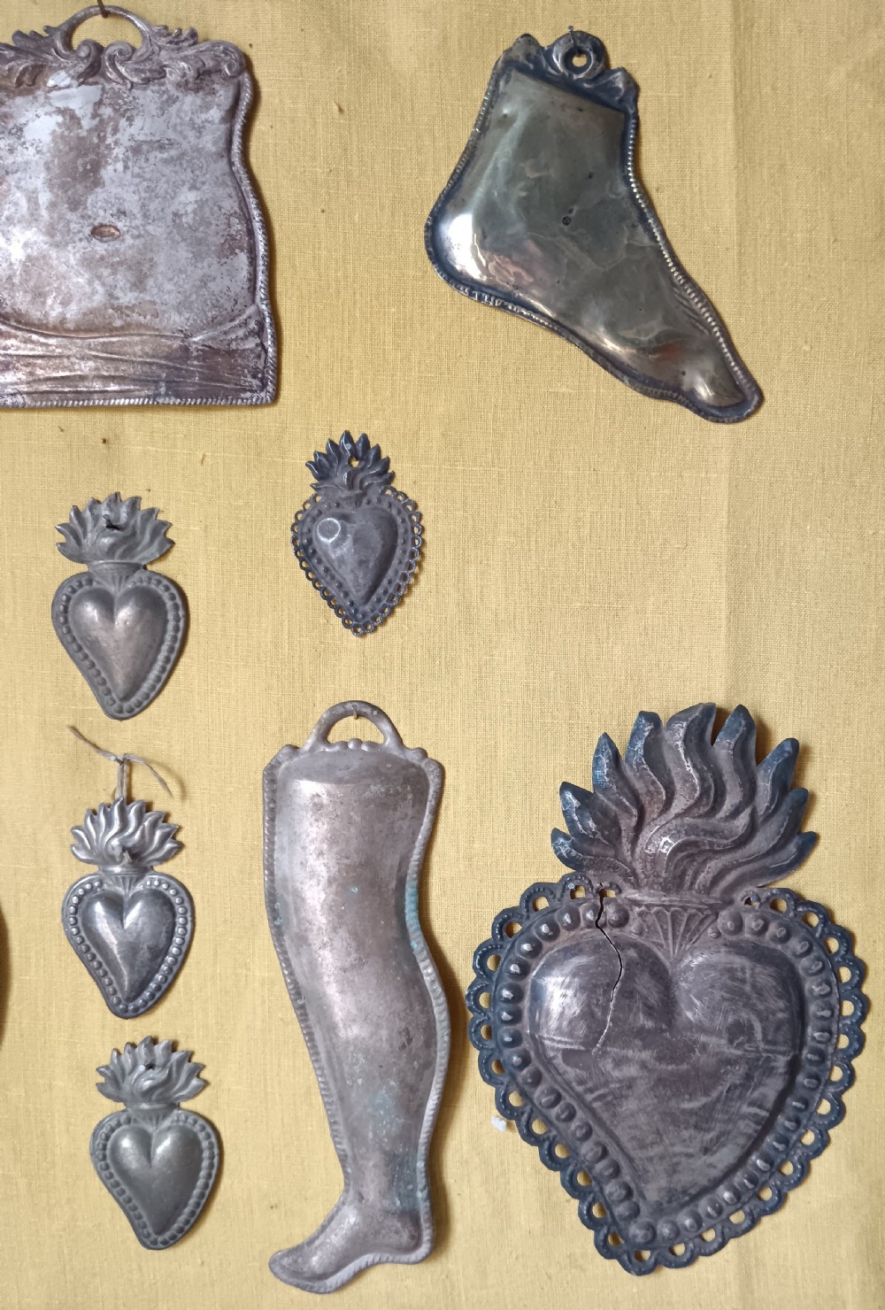 Seventeen silver-coloured metal ex-voto tokens arranged on yellow fabric inside a dark wooden frame. View VI