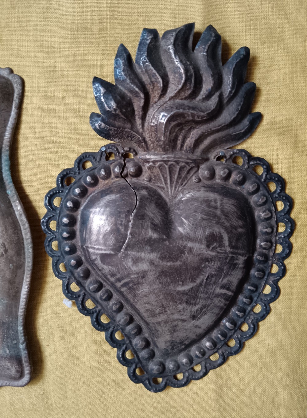 Seventeen silver-coloured metal ex-voto tokens arranged on yellow fabric inside a dark wooden frame. View VII