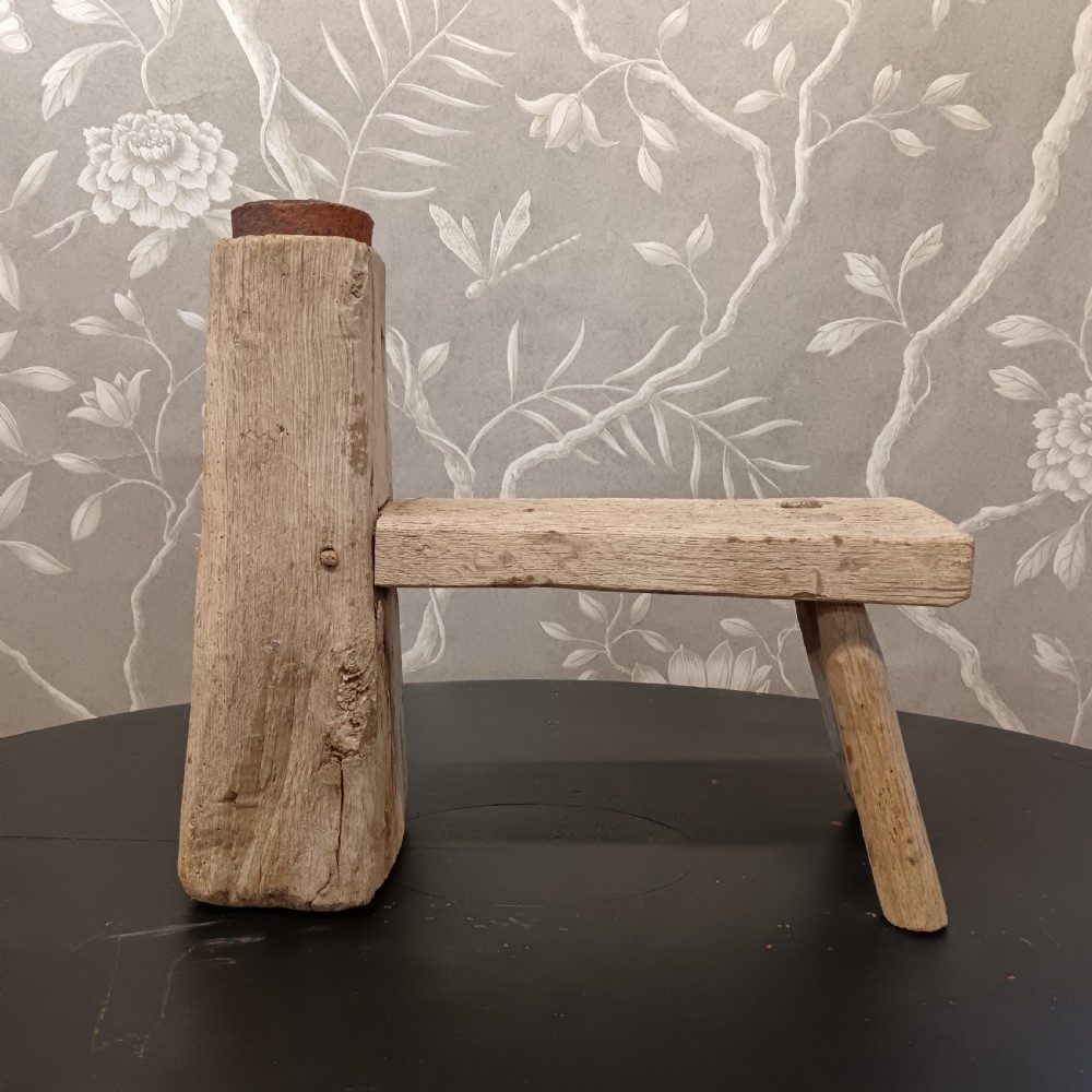 An early 19th-century wooden cobbler stool with a thick vertical timber post topped with iron, joined to a low horizontal bench surface with splayed legs.