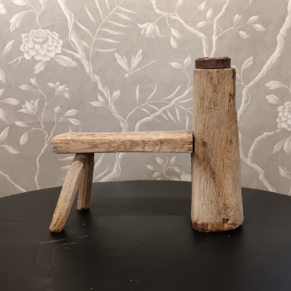An early 19th-century wooden cobbler stool with a thick vertical timber post topped with iron, joined to a low horizontal bench surface with splayed legs. View II