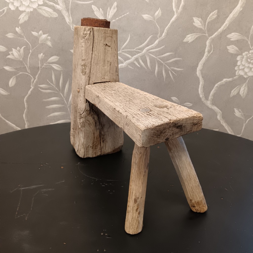 An early 19th-century wooden cobbler stool with a thick vertical timber post topped with iron, joined to a low horizontal bench surface with splayed legs. View VI