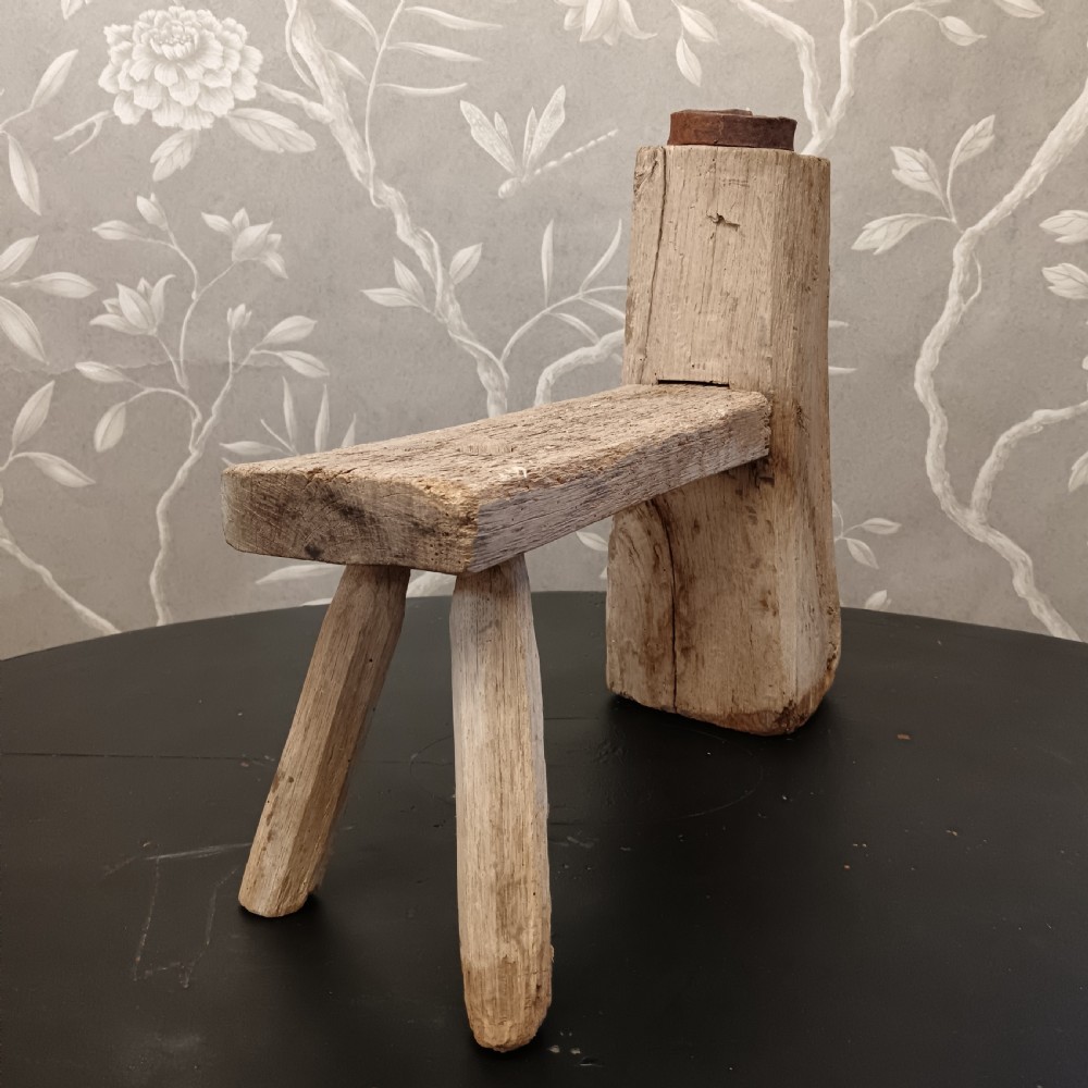 An early 19th-century wooden cobbler stool with a thick vertical timber post topped with iron, joined to a low horizontal bench surface with splayed legs. View VII