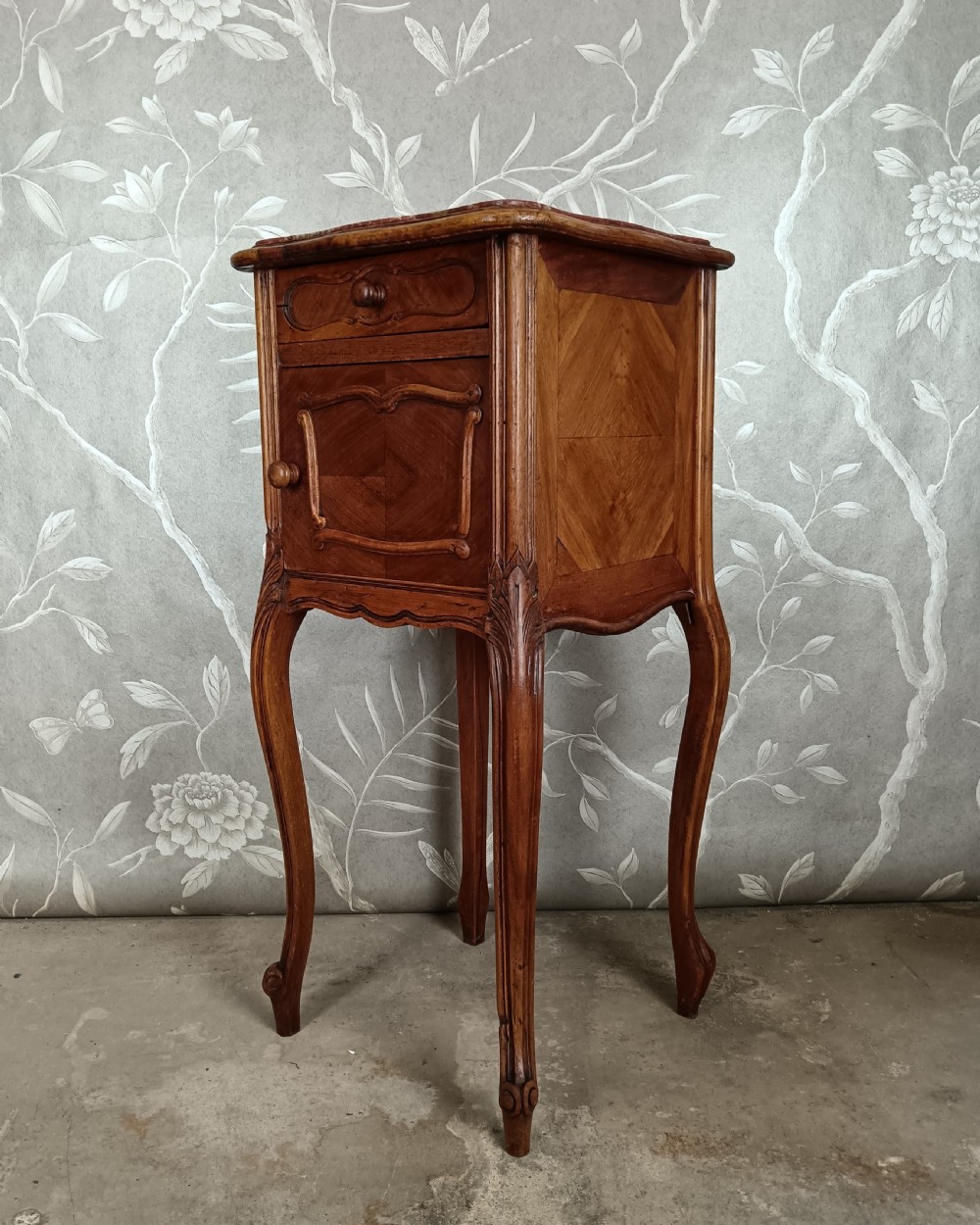 A tall French bedside cupboard crafted from warm brown wood with slim cabriole legs, parquetry panels, a single top drawer, and a lower cabinet door.