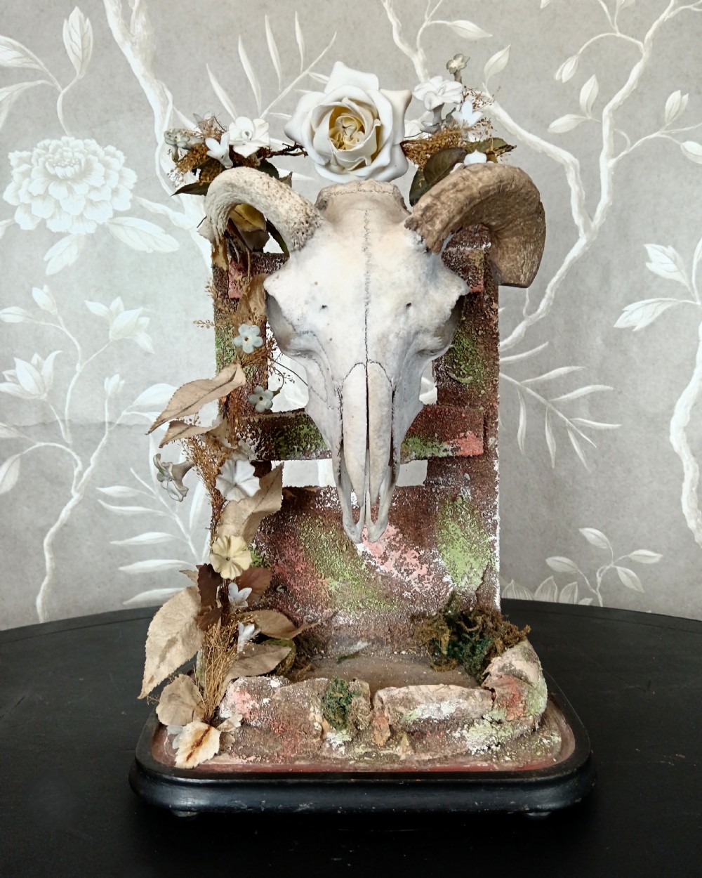 An antique Victorian animal skull with curved horns mounted on a moss-covered diorama base decorated with dried flowers, set upon a dark oval wooden plinth.