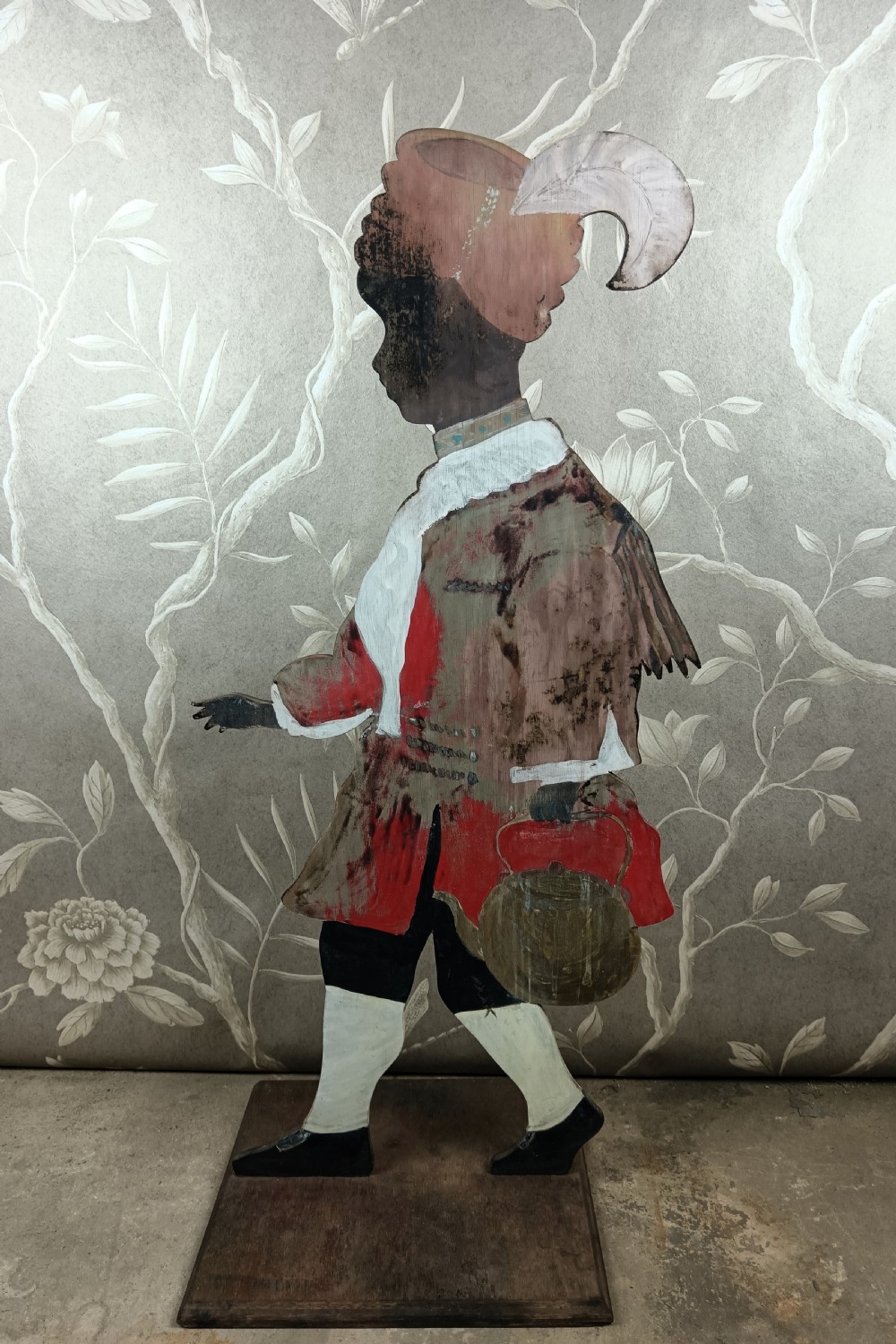 An early twentieth-century painted wooden dummy board figure depicting a man in a red coat and feathered hat holding a kettle.