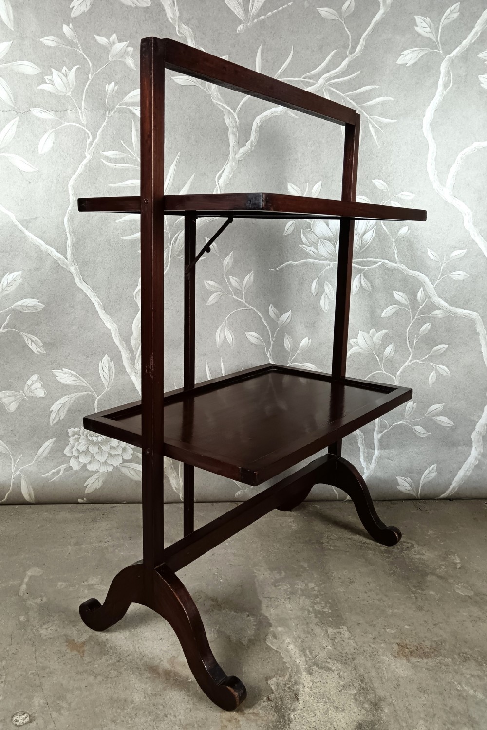 A dark mahogany two-tier folding dumb waiter with rectangular tray surfaces, upper carrying handle, and splayed legs. View V