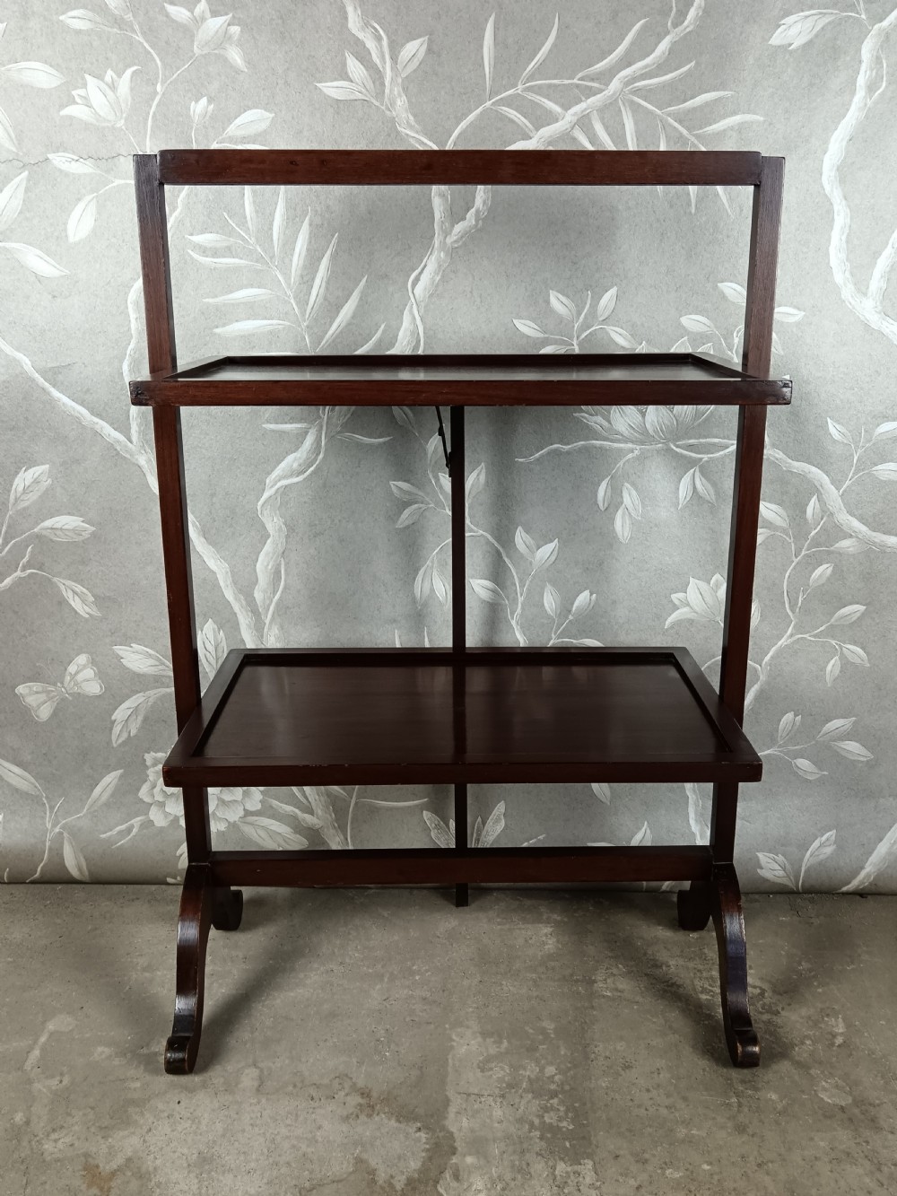 A dark mahogany two-tier folding dumb waiter with rectangular tray surfaces, upper carrying handle, and splayed legs.