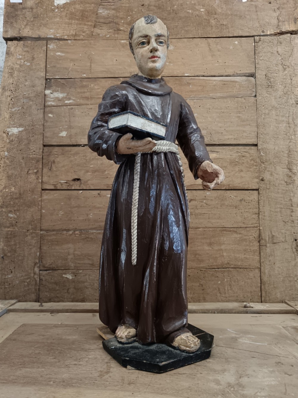 An antique carved wooden figure depicting Saint Anthony wearing a dark brown habit and holding a book.