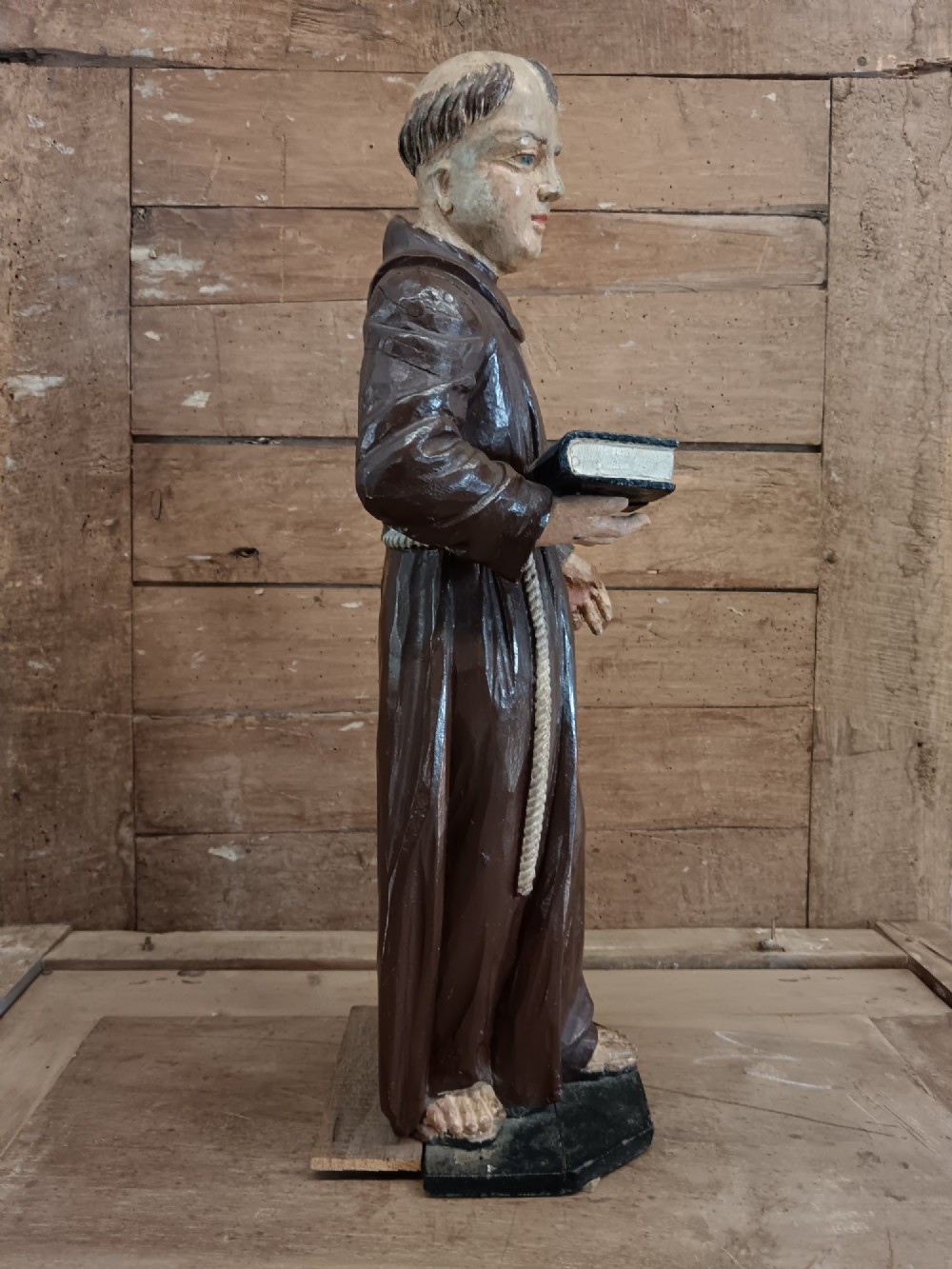 An antique carved wooden figure depicting Saint Anthony wearing a dark brown habit and holding a book. View X