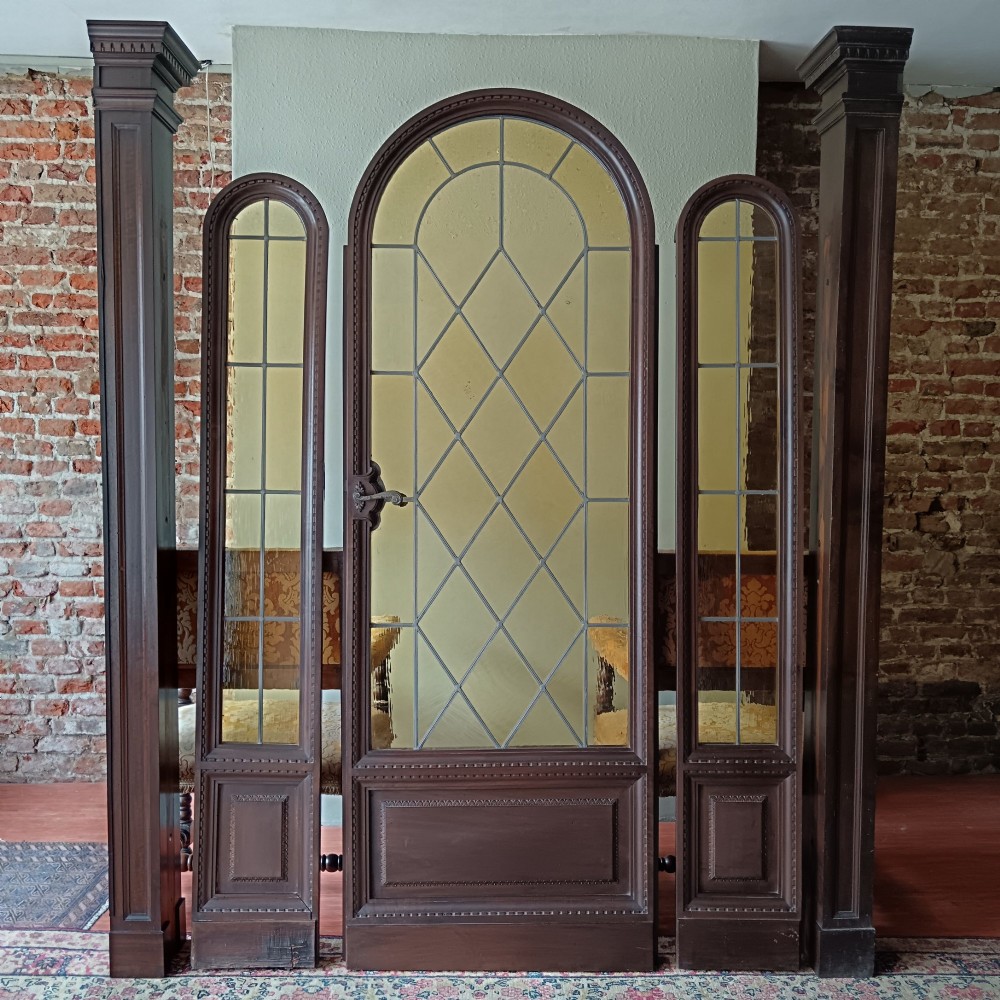 An antique dark wood vestibule doorway featuring a central arched leaded glass door flanked by two slender side lights and classic fluted columns.