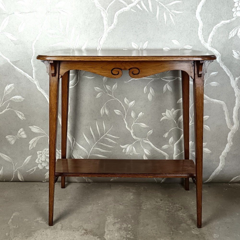 An antique dark oak two-tier side table featuring a carved scroll motif on the apron and tapered square legs.
