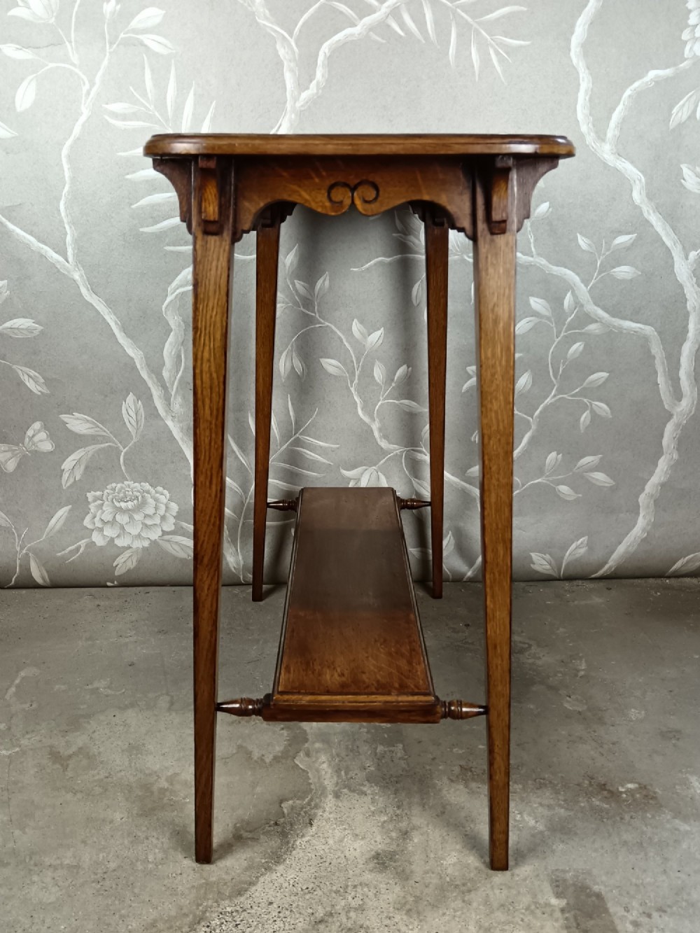 An antique dark oak two-tier side table featuring a carved scroll motif on the apron and tapered square legs. View II