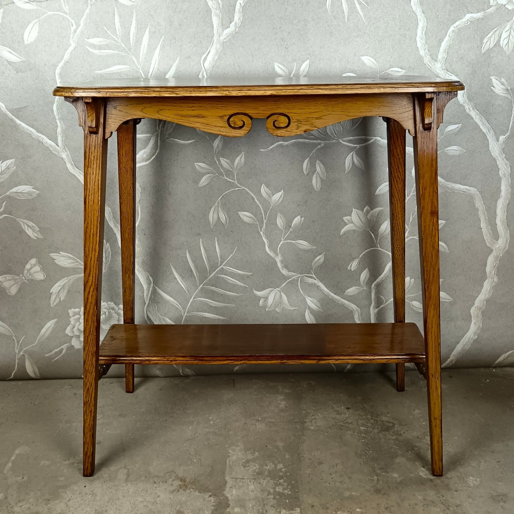 An antique dark oak two-tier side table featuring a carved scroll motif on the apron and tapered square legs. View III