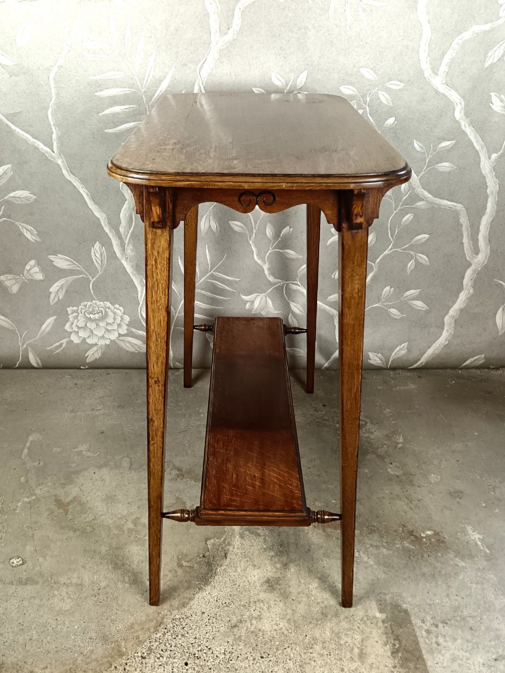 An antique dark oak two-tier side table featuring a carved scroll motif on the apron and tapered square legs. View IV