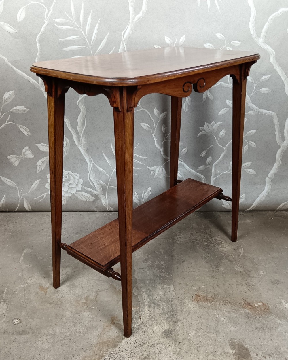 An antique dark oak two-tier side table featuring a carved scroll motif on the apron and tapered square legs. View V