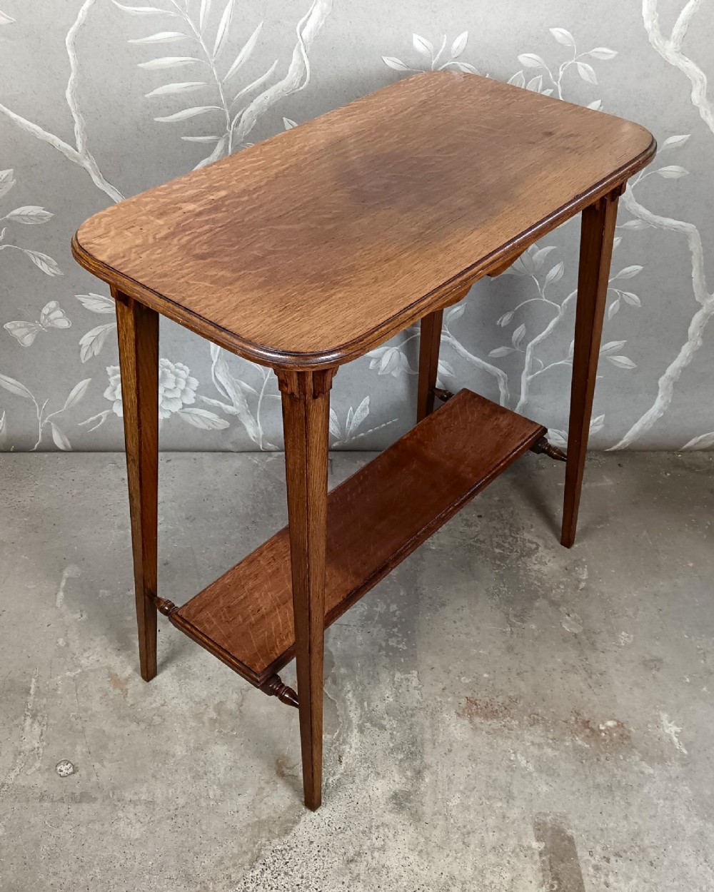 An antique dark oak two-tier side table featuring a carved scroll motif on the apron and tapered square legs. View VI