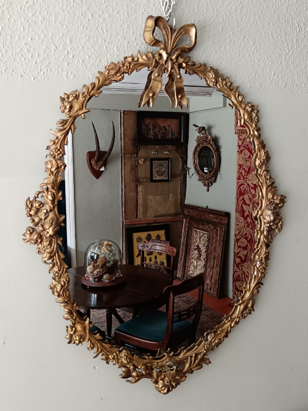 An oval gilt bronze wall mirror with a detailed floral foliate frame and a ribbon bow cresting at the top.