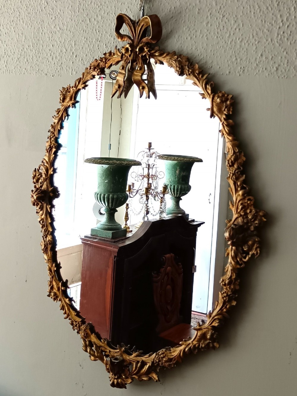 An oval gilt bronze wall mirror with a detailed floral foliate frame and a ribbon bow cresting at the top. View II