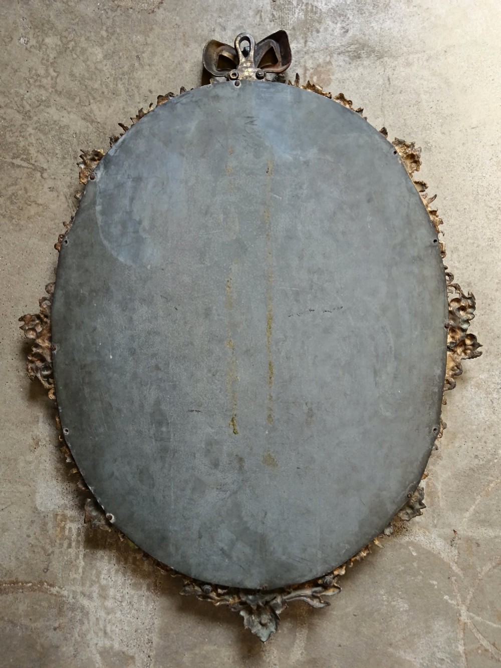 An oval gilt bronze wall mirror with a detailed floral foliate frame and a ribbon bow cresting at the top. View V