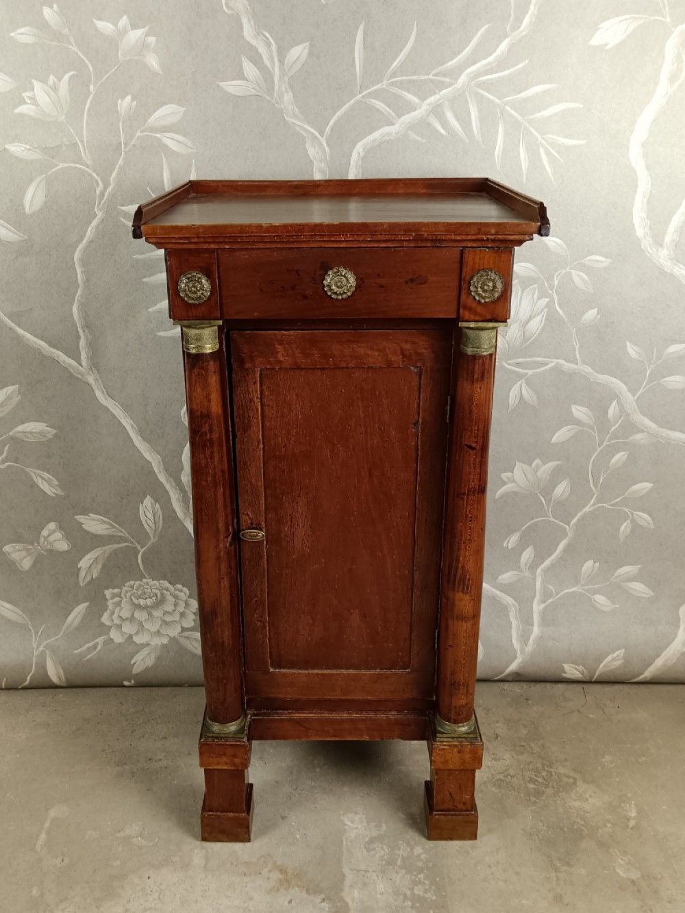 A nineteenth-century Italian Empire walnut bedside cabinet featuring a top drawer, single panelled door, brass rosette mounts, and flanking column columns.