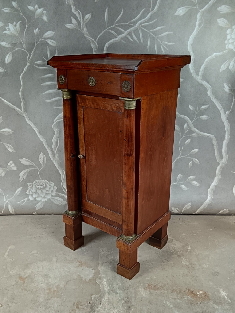 A nineteenth-century Italian Empire walnut bedside cabinet featuring a top drawer, single panelled door, brass rosette mounts, and flanking column columns. View II