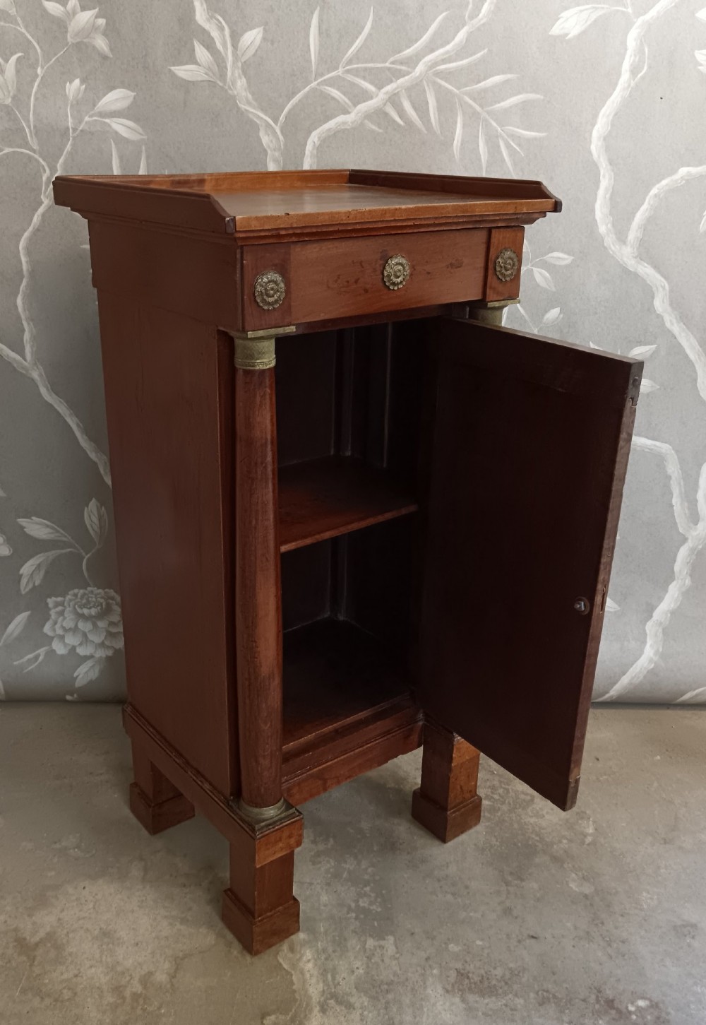 A nineteenth-century Italian Empire walnut bedside cabinet featuring a top drawer, single panelled door, brass rosette mounts, and flanking column columns. View IV