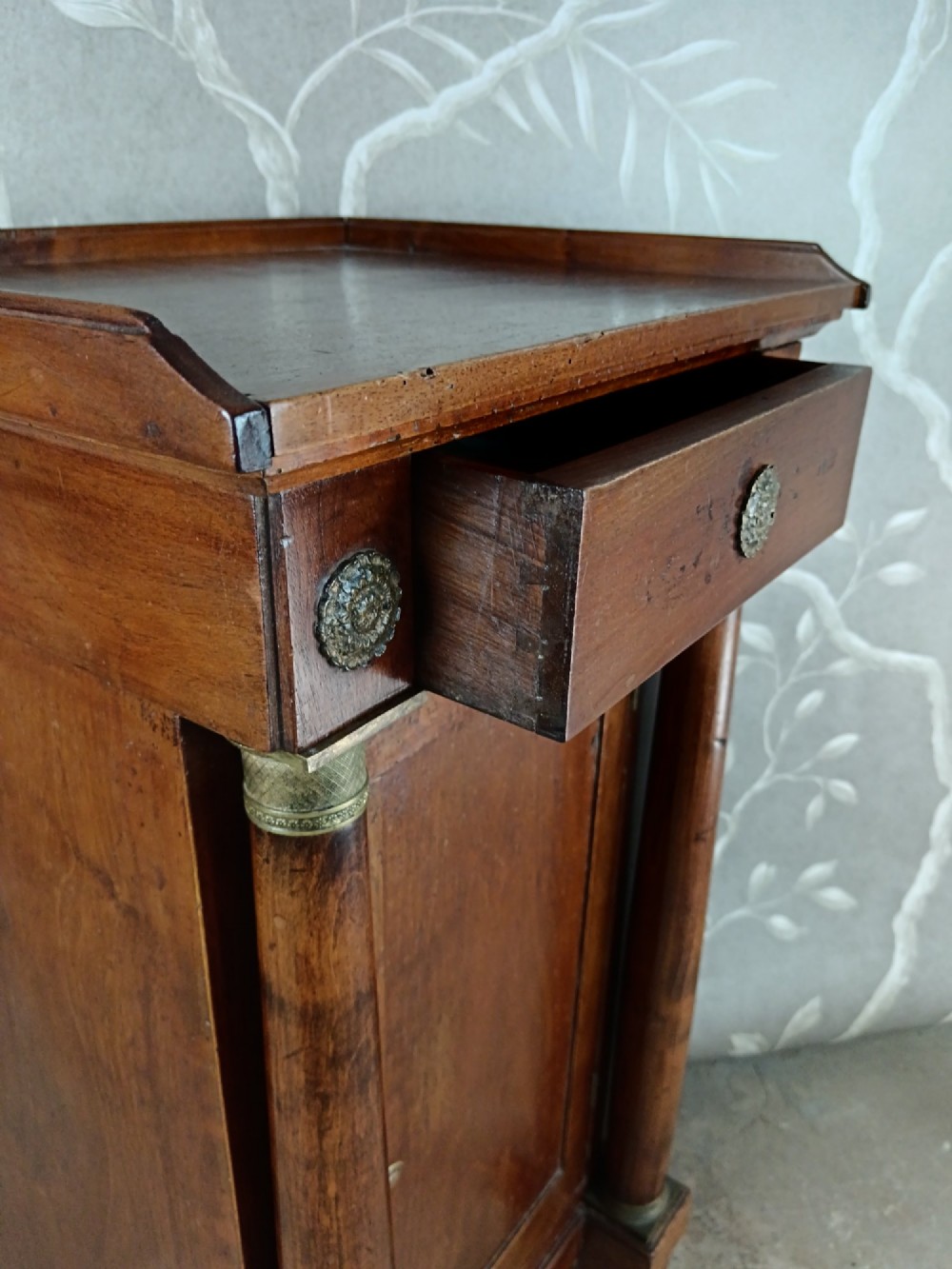 A nineteenth-century Italian Empire walnut bedside cabinet featuring a top drawer, single panelled door, brass rosette mounts, and flanking column columns. View VI