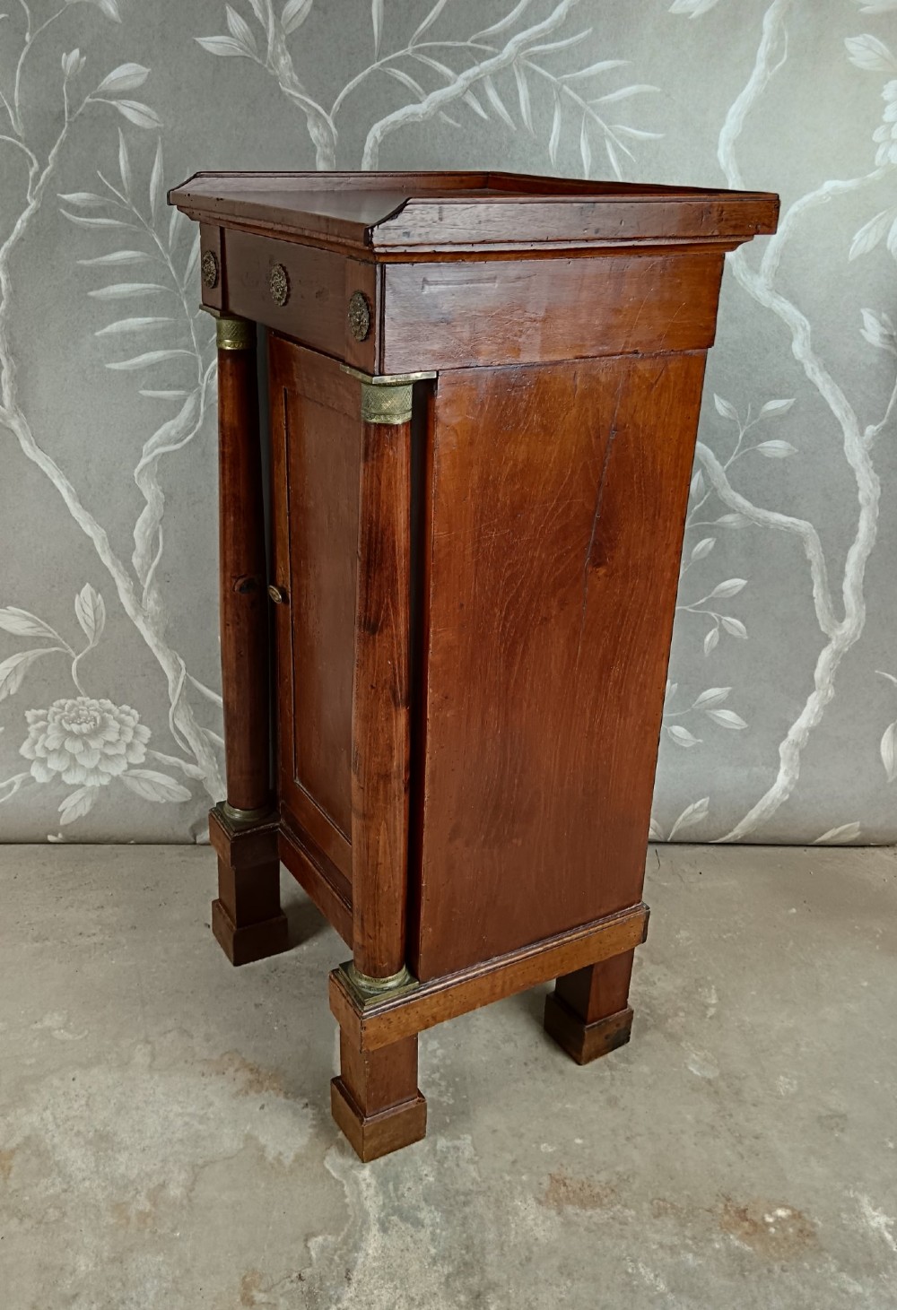 A nineteenth-century Italian Empire walnut bedside cabinet featuring a top drawer, single panelled door, brass rosette mounts, and flanking column columns. View VIII