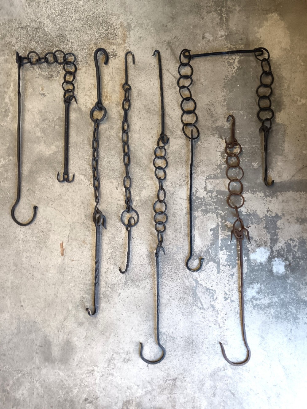 A collection of six hand-forged dark wrought iron adjustable chimney trammel hooks and pot hangers laid flat on a concrete background.
