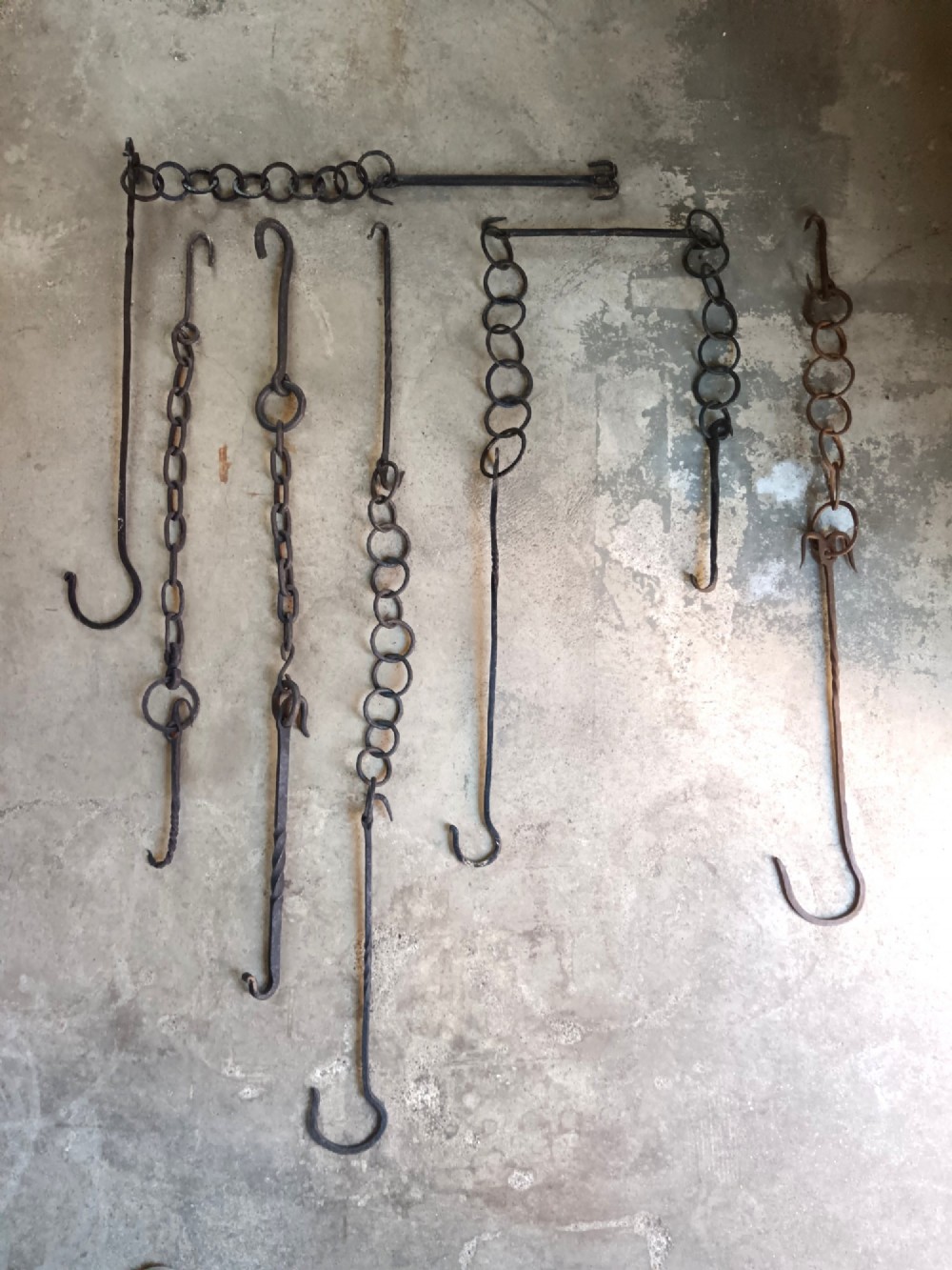 A collection of six hand-forged dark wrought iron adjustable chimney trammel hooks and pot hangers laid flat on a concrete background. View II