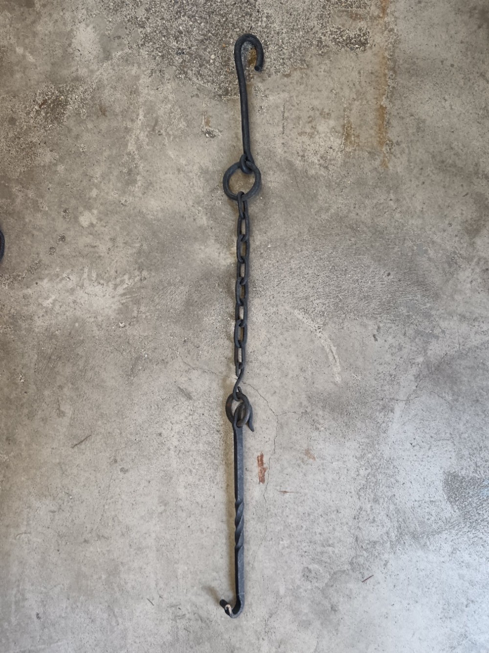 A collection of six hand-forged dark wrought iron adjustable chimney trammel hooks and pot hangers laid flat on a concrete background. View III