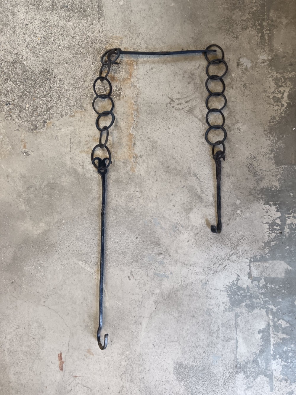 A collection of six hand-forged dark wrought iron adjustable chimney trammel hooks and pot hangers laid flat on a concrete background. View IV