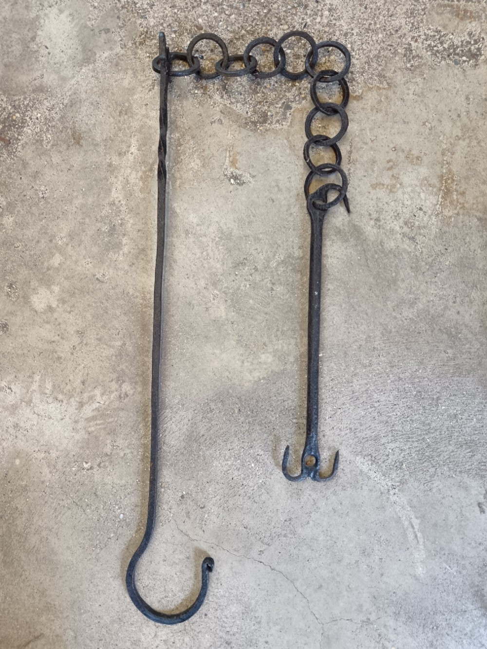 A collection of six hand-forged dark wrought iron adjustable chimney trammel hooks and pot hangers laid flat on a concrete background. View V