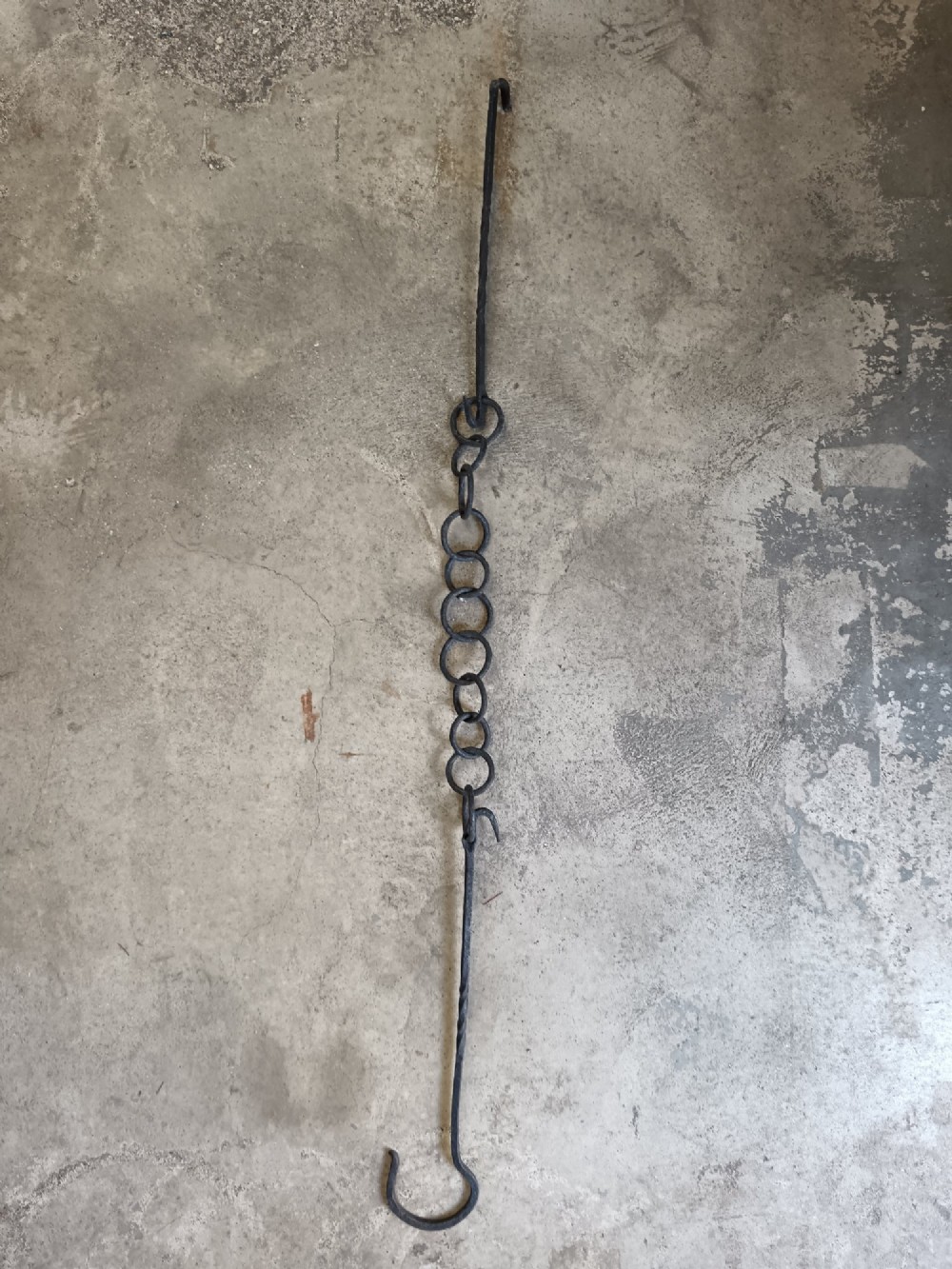 A collection of six hand-forged dark wrought iron adjustable chimney trammel hooks and pot hangers laid flat on a concrete background. View VI