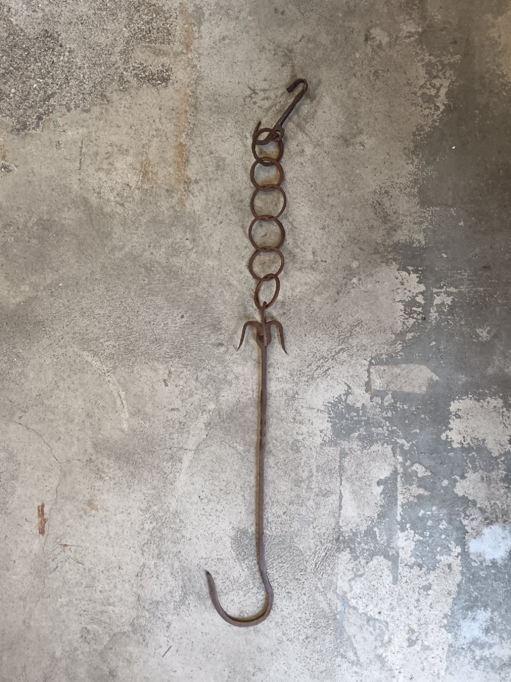 A collection of six hand-forged dark wrought iron adjustable chimney trammel hooks and pot hangers laid flat on a concrete background. View VII